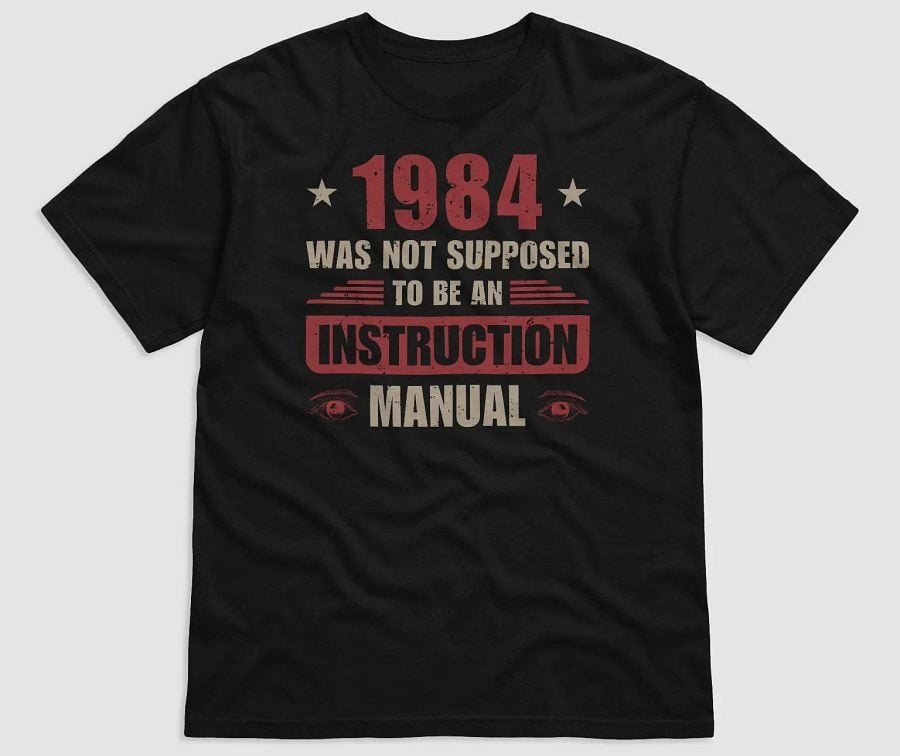 Black t-shirt with printed text in red and beige reading '1984 was not supposed to be an instruction manual' accompanied by two stars and stylized eyes below the text.